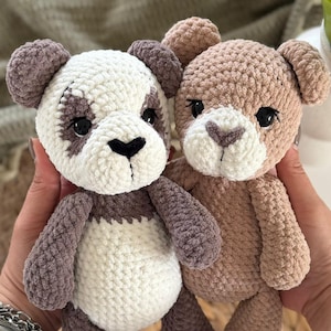 Könnte beinhalten: Zwei handgefertigte Häkelteddybären, einer als Panda. Der Pandabär hat einen weißen Körper mit lila und schwarzen Akzenten. Der andere ist hellbraun. Beide haben schwarze Augen und werden in Händen gehalten.