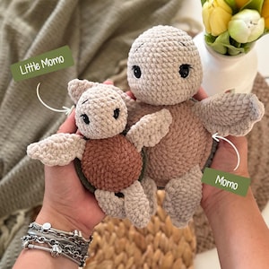 Op de afbeelding: Twee gehaakte schildpad knuffels gemaakt van zacht, beige en bruin garen. De kleinere schildpad is gelabeld als Little Momo en de grotere als Momo. Beide speeltjes hebben ronde koppen, donkere plastic ogen en een textuurrijk lichaam.