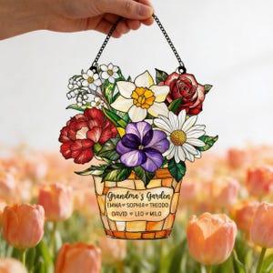 May include: A hanging floral decoration featuring a bouquet of colorful flowers in a mosaic-style pot. The flowers include red roses, white daisies, and purple pansies. The text "Grandma's Garden" is displayed on the pot, along with names.