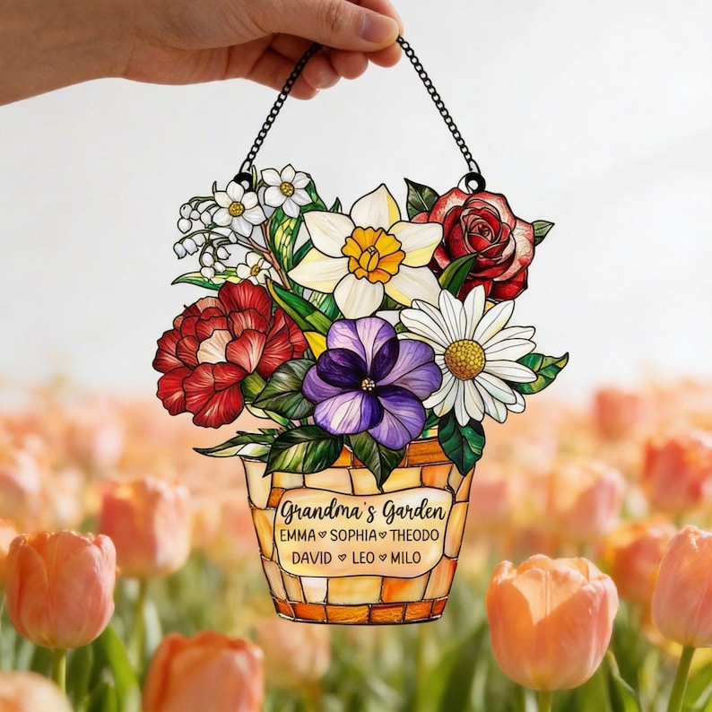 May include: A hanging floral decoration featuring a bouquet of colorful flowers in a mosaic-style pot. The flowers include red roses, white daisies, and purple pansies. The text "Grandma's Garden" is displayed on the pot, along with names.