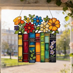 May include: Stained glass window decoration featuring a colorful design of books and flowers. The flowers include orange, yellow, red, blue, and white blooms. The books are various colors, including yellow, red, blue, and green. The window hanging is suspended by a clear string.