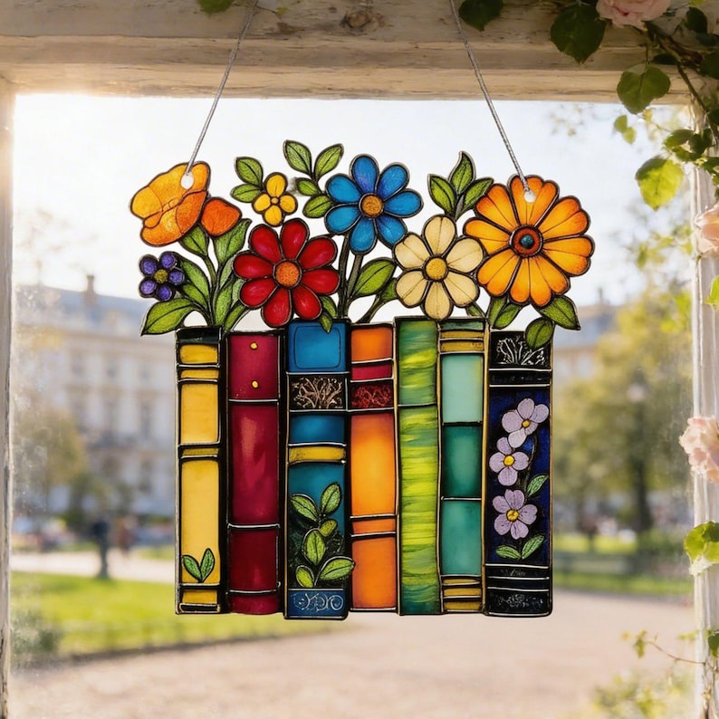 May include: Stained glass window decoration featuring a colorful design of books and flowers. The flowers include orange, yellow, red, blue, and white blooms. The books are various colors, including yellow, red, blue, and green. The window hanging is suspended by a clear string.