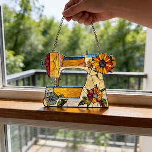 May include: A stained glass sewing machine suncatcher, designed for hanging. The artwork features a vibrant palette of yellow, orange, and blue. It is adorned with floral details, including sunflowers and other flowers. A decorative window ornament.