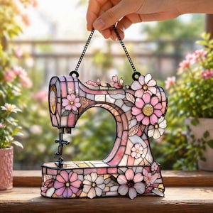 May include: A decorative stained glass sewing machine in pink, white, and brown, with floral embellishments. It hangs from a black chain and features a detailed needle and thread area. The design uses a mosaic pattern. The piece is suitable for a window.