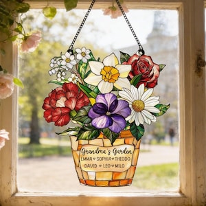 May include: A stained glass window hanging featuring a floral arrangement in a decorative pot. The flowers include red roses, white daisies, yellow daffodils, and purple pansies. The pot is inscribed with "Grandma's Garden" and several names. The window is suspended by a black chain.