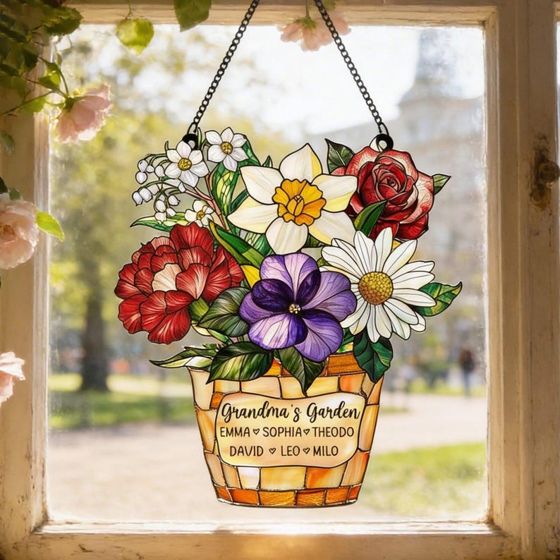May include: A stained glass window hanging featuring a floral arrangement in a decorative pot. The flowers include red roses, white daisies, yellow daffodils, and purple pansies. The pot is inscribed with "Grandma's Garden" and several names. The window is suspended by a black chain.