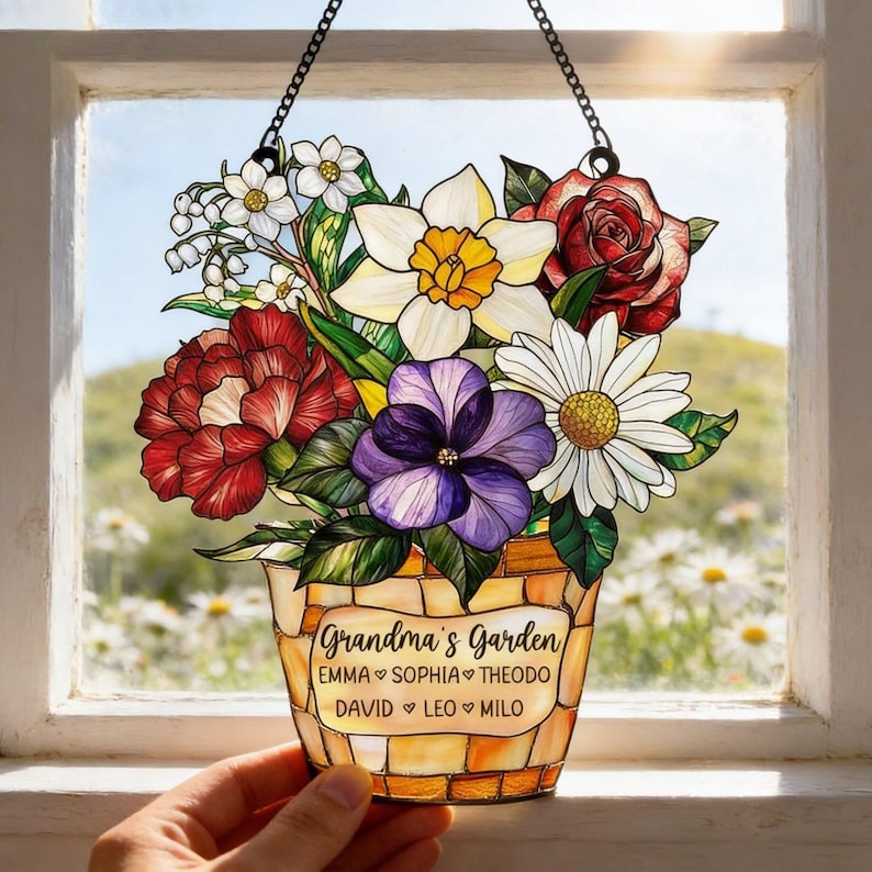 May include: Stained glass floral bouquet in a decorative pot, hanging from a chain. The bouquet includes red roses, white daisies, and purple pansies. The pot has the words "Grandma's Garden" and names below. The background is a window with a field of daisies.
