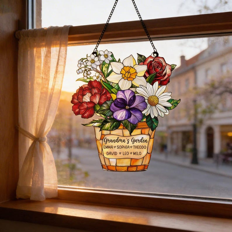 May include: A stained glass window hanging featuring a basket of colorful flowers. The basket is filled with red, white, purple, and yellow flowers. The text "Grandma's Garden" is displayed on the basket, along with names.