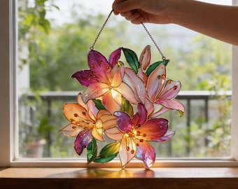 May include: Stained glass floral art piece featuring lilies in shades of pink, orange, and purple, with green leaves. The artwork is held by a chain and hangs in front of a window, showcasing the vibrant colors and intricate details of the design.