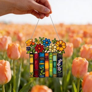 May include: A colorful stained glass panel featuring a row of books topped with vibrant flowers. The flowers include orange, red, blue, and yellow blooms. The panel is held by a silver wire, and the background is a field of orange tulips.