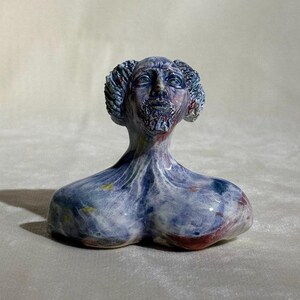 May include: A ceramic sculpture of a stylized human head and torso. The sculpture is primarily blue with accents of red, yellow, and white. The head has curly hair and a beard. The torso has a rounded shape.