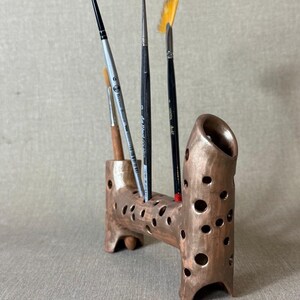 May include: A handmade, brown ceramic brush holder with multiple holes and a unique, organic shape. Several paint brushes of varying sizes and shapes are displayed in the holder, with yellow, black, and silver handles.