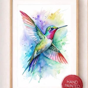 Pode incluir: Uma pintura em aquarela emoldurada de um beija-flor em voo. O pássaro apresenta cores vibrantes, incluindo verde, rosa, azul e amarelo. O fundo mostra uma lavagem em aquarela colorida. Um adesivo redondo vermelho com o texto "HAND PAINTED" é visível.