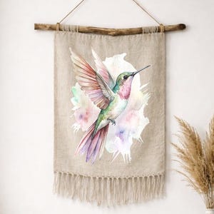 Może przedstawiać: Wiszący obraz ścienny z kolibrem. Akwarelowy wzór przedstawia kolibra w odcieniach różu, zieleni i fioletu na miękkim, kolorowym tle. Obraz ścienny wykonany jest z tkaniny w naturalnym kolorze z frędzlami.