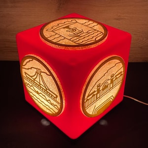 Può includere: Una lampada rossa a forma di cubo con disegni ovali illuminati su ogni lato. I disegni sembrano essere di ponti ed edifici. La lampada è collegata e emette un bagliore caldo.