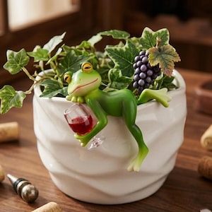 May include: A white ceramic planter with a wavy texture containing green ivy and a cluster of purple grapes. A green frog figurine is attached to the rim, holding a small clear glass filled with red liquid, appearing to lounge on the edge.