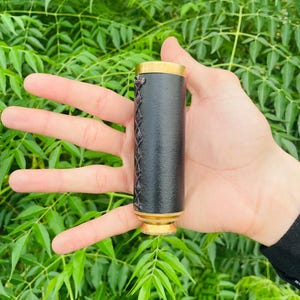 Peut inclure: Un télescope monoculaire cylindrique portatif doté d'un revêtement en cuir noir texturé avec des détails en point de croix marron foncé sur le côté, rehaussé par des bords en métal doré poli aux extrémités supérieure et inférieure.