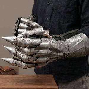 May include: A metallic gauntlet featuring articulated finger plates with pointed tips and intricate floral engravings. The armor piece is constructed from polished steel segments connected by rivets, covering the hand and wrist with a protective cuff.