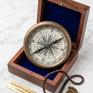 May include: A brass navigation compass with a white dial and black cardinal markings sits inside a dark wooden box lined with blue velvet. A string of brown wooden beads and a small spiral ammonite fossil are placed next to the box.