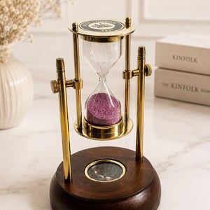 Puede incluir: Un reloj de arena de latón con arena morada, montado sobre un marco giratorio encima de una base circular de madera marrón oscuro que contiene una brújula. La parte superior del reloj tiene una etiqueta negra con el texto ROORKEE LONDON 1917.