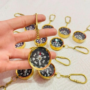 Può includere: Una collezione di portachiavi a bussola in metallo dorato con cassa circolare, quadrante nero, indicazioni cardinali bianche e un ago con punta rossa. Ogni bussola è fissata a una catenella corta e a un moschettone in metallo.