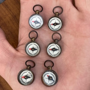Puede incluir: Seis pequeñas brújulas de estilo antiguo con acabado bronce. Cada brújula presenta una esfera blanca con marcas direccionales negras y rojas y un pequeño lazo en la parte superior. Las brújulas están dispuestas sobre una superficie marrón claro.