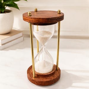 May include: An hourglass featuring a clear glass bulb filled with white sand, held between two circular dark wood bases. The structure is supported by three vertical gold-toned metal rods, with small gold-toned decorative knobs on the top base.