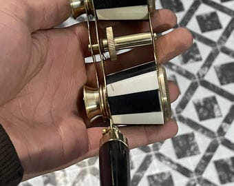 May include: Opera glasses featuring black and white striped barrels with brass accents and a central focusing wheel. A dark, curved handle hangs from the base. The text Thecuriositycabgifts is visible in the top right corner.