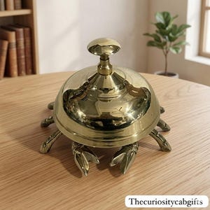 Può includere: Un campanello da scrivania in ottone lucido a forma di granchio, caratterizzato da una cupola arrotondata con una maniglia centrale e molteplici zampe articolate che si estendono dalla base. Il testo Thecuriositycabgifts è nell'angolo in basso.