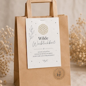 Könnte beinhalten: Eine braune Papiertüte mit einer rechteckigen weißen Karte auf der Vorderseite. Die Karte zeigt ein goldenes Blume des Lebens-Symbol, eine Strichzeichnung einer Pflanze und den Text Wilde Weiblichkeit, Kleine Auszeiten für deine weibliche Kraft, Selbstliebe und Verbundenheit.