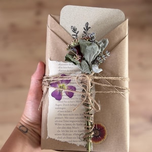 Könnte beinhalten: Ein rechteckiges Paket in braunem Papier, umwickelt mit Paketschnur und verziert mit einem kleinen Trockenblumenstrauß, einer gepressten violetten Blume und einem Stück einer Buchseite mit deutschem Text. Ein kleiner Blumenaufkleber ist vorne.