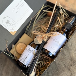 Könnte beinhalten: Eine schwarze Geschenkbox mit zwei braunen Glasflaschen mit weißen Etiketten, einer Sprühflasche und einem Schilfrohrdiffusor. Die Flaschen sind mit Bindfaden und einer hölzernen Eichel gebunden. Die Box enthält auch Holzscheiben und getrocknetes Gras.