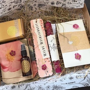 Könnte beinhalten: Eine Geschenkbox mit verschiedenen Artikeln, darunter eine braune Glas-Sprühflasche mit goldenem geometrischen Design, ein rosa Lesezeichen mit dem Text "WILDE WEIBLICHKEIT" und eine kleine Box mit goldenem Design. Die Box ist mit floral gemustertem Papier ausgekleidet.