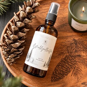 Könnte beinhalten: Eine braune Glassprühflasche mit einem weißen Etikett mit der Aufschrift Waldgeflüster und Natur Raumspray, begleitet von einem Tannenzapfen und einer grünen Kerze auf einem Holztablett mit lasergraviertem Tannenzapfen-Motiv.