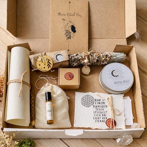 Könnte beinhalten: Eine Moon Ritual Box mit spirituellen Gegenständen. Enthält eine gerollte Pergamentrolle, eine kleine Flasche, ein Salbeibündel, ein Mond-Ornament aus Holz und weitere Artikel. Die Pappschachtel enthält eine Karte mit dem Text "Moon Ritual Box".