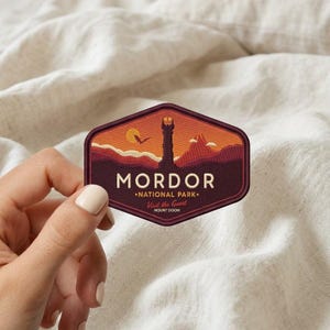 May include: A hexagonal embroidered patch featuring a landscape of Mordor with a central dark tower, a fiery mountain, and a yellow sun. The text reads MORDOR NATIONAL PARK, Visit the Great, and MOUNT DOOM in cream and red lettering on a dark background.