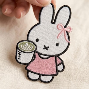 May include: An embroidered patch featuring a white rabbit character with long ears, wearing a pink dress and a small pink bow on its head. The rabbit is holding a white mug filled with a green latte featuring white foam art, outlined in black thread.