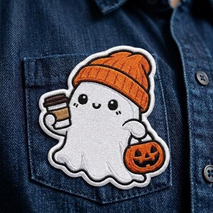 May include: An embroidered patch featuring a white ghost wearing an orange beanie, holding a brown coffee cup in one hand and an orange jack-o'-lantern pumpkin bucket in the other, with black eyes and a small smile on a white background.
