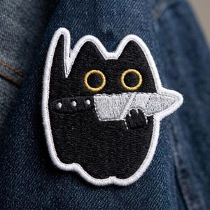 May include: An embroidered patch featuring a black cat with large yellow eyes and a white border. The cat is depicted holding a silver-colored knife in its mouth, with a black collar featuring small silver studs visible around its neck area.