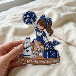 May include: An embroidered patch featuring a cheerleader with brown hair and a blue and white uniform, holding a pom-pom. It includes a megaphone labeled Isabella, a duffel bag, and a base with the text Just A Girl Who Loves Cheerleading.