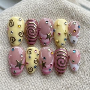 May include: A set of ten artificial nails with a variety of designs. Some nails are pale yellow with gold swirls and rhinestones. Others are pink with gold stars and stripes. The remaining nails have a pink and white design with rhinestones.