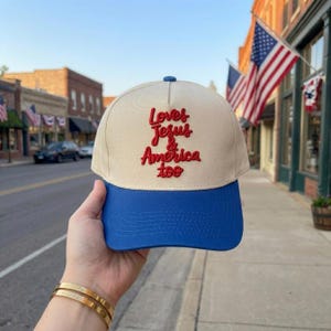 May include: A beige and blue baseball cap with the embroidered text "Loves Jesus & America too" in red. The cap has a blue brim and a blue button on top. The image is taken outdoors with American flags in the background.