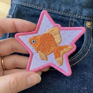 May include: An embroidered patch in the shape of a five-pointed star with a light purple center and a bright pink border. Centered on the star is a detailed orange goldfish with textured scales, a white eye with a black pupil, and tan fins.