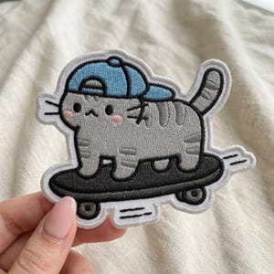 May include: An embroidered patch featuring a gray tabby cat with pink cheeks wearing a light blue baseball cap. The cat is standing on a black skateboard with dark gray wheels. The design has a white border and includes small motion lines.