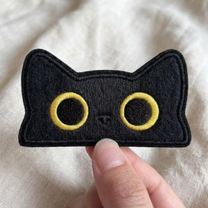 May include: A black embroidered patch shaped like a cat head with two pointed ears. The design features two large, circular eyes with bright yellow outlines and black centers, along with a small, simple stitched nose detail in the center.