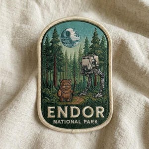 May include: An embroidered patch with a cream border featuring a forest scene with a brown Ewok, a grey AT-ST walker, and a Death Star in the sky. The text ENDOR NATIONAL PARK is stitched in white at the bottom against a dark green background.