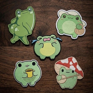 May include: Five embroidered patches featuring green frogs in various poses: one standing, one with a hat and basket, one crying, one holding a yellow mug, and one wearing a red and white mushroom cap while holding a small stringed instrument.