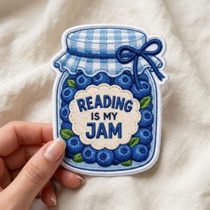 May include: An embroidered patch shaped like a glass jar filled with blueberries. The jar lid has a blue and white gingham pattern with a blue bow. A cream-colored scalloped label in the center features the dark blue text READING IS MY JAM.