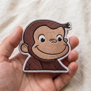 May include: An embroidered iron-on patch featuring the character Curious George. The design shows the monkey's head and shoulders in shades of dark and light brown thread, with large white eyes, black pupils, and a wide smile, outlined in white.