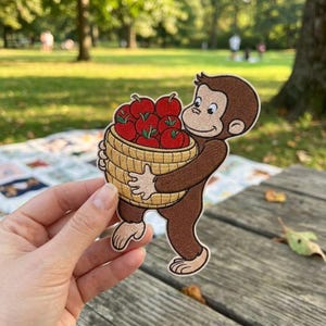 May include: An embroidered patch featuring a brown monkey holding a tan woven basket filled with bright red apples with green stems. The monkey has a light tan face, ears, and feet, with a dark brown body and a smiling expression.